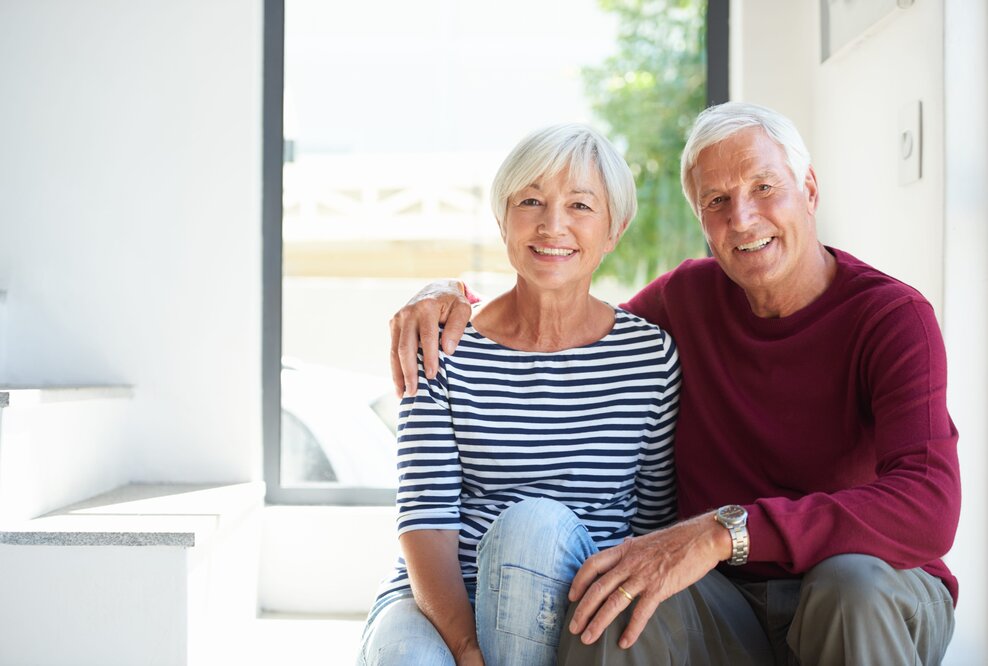 Lächelndes Seniorenpaar sitzt im Tageslicht vor einem Fenster seines Eigenheims nach Immobilien Teilverkauf.