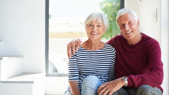 Lächelndes Seniorenpaar sitzt im Tageslicht vor einem Fenster seines Eigenheims nach Immobilien Teilverkauf.