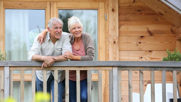 Älteres Paar steht lächelnd auf Balkon mit Holzfassade im Hintergrund.